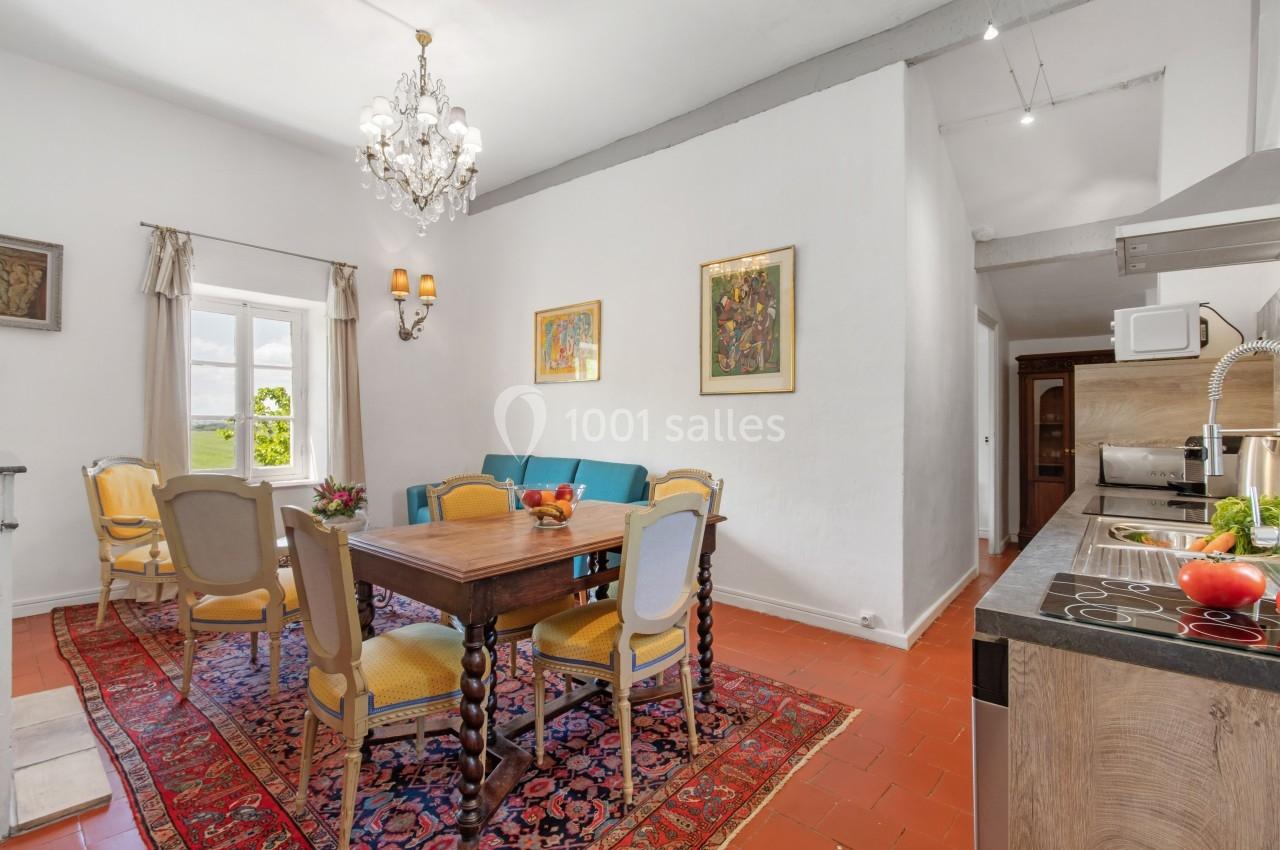 Salle à manger lumineuse avec table en bois, chaises jaunes, tapis coloré et cuisine ouverte en arrière-plan.