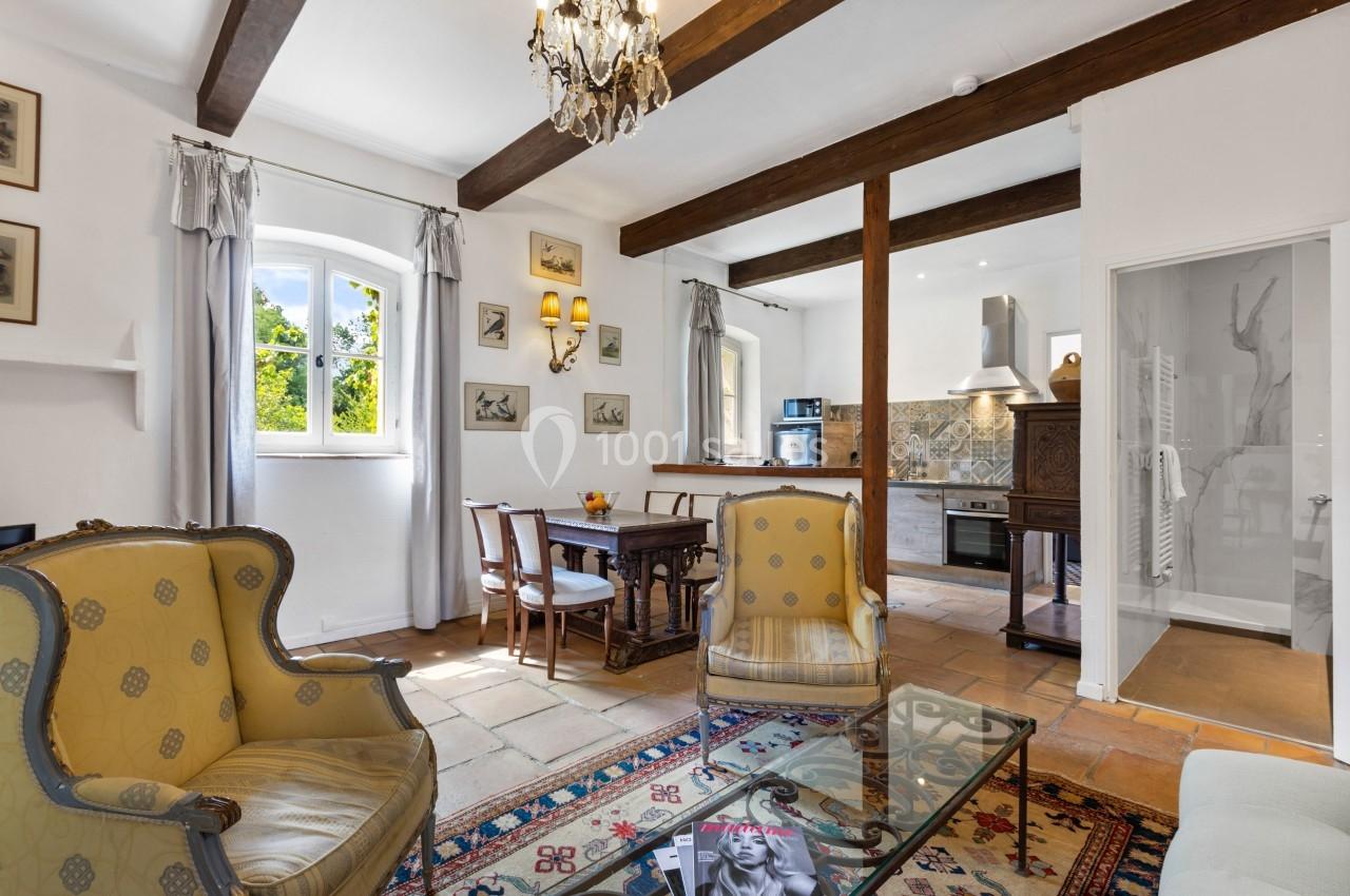 Salon lumineux avec poutres apparentes, meubles anciens, table à manger, et vue sur une cuisine ouverte.