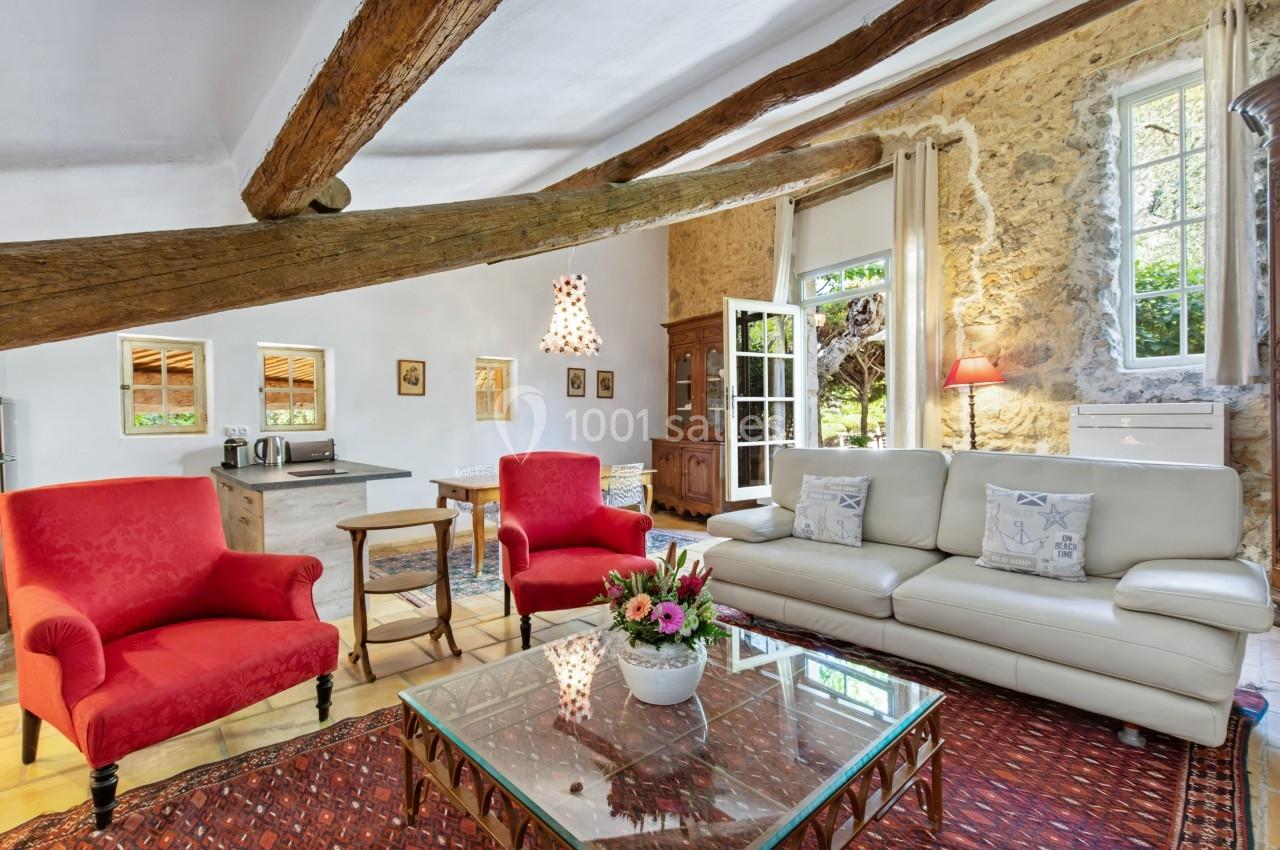 Salon lumineux avec poutres apparentes, canapé blanc, fauteuils rouges, table basse en verre et murs en pierre.