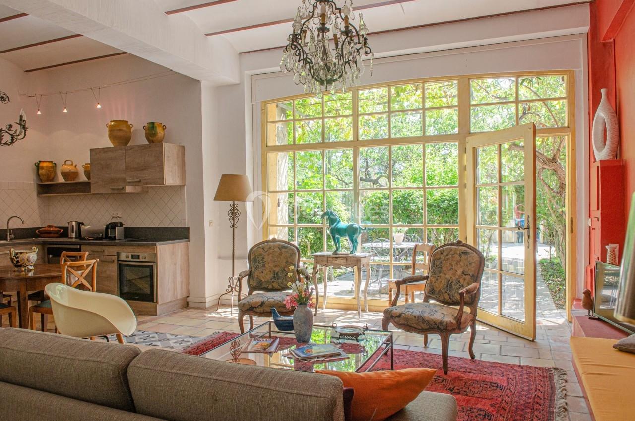 Salon lumineux avec grandes baies vitrées ouvertes sur un jardin, mobilier vintage et cuisine ouverte en arrière-plan.