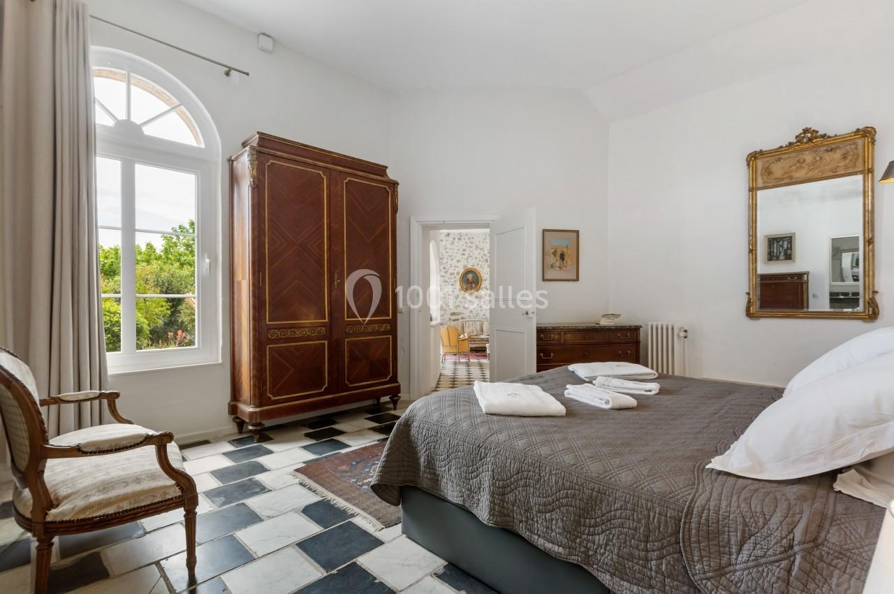 Chambre lumineuse avec lit double, armoire en bois, miroir doré, fenêtre cintrée et sol en carreaux noirs et blancs.