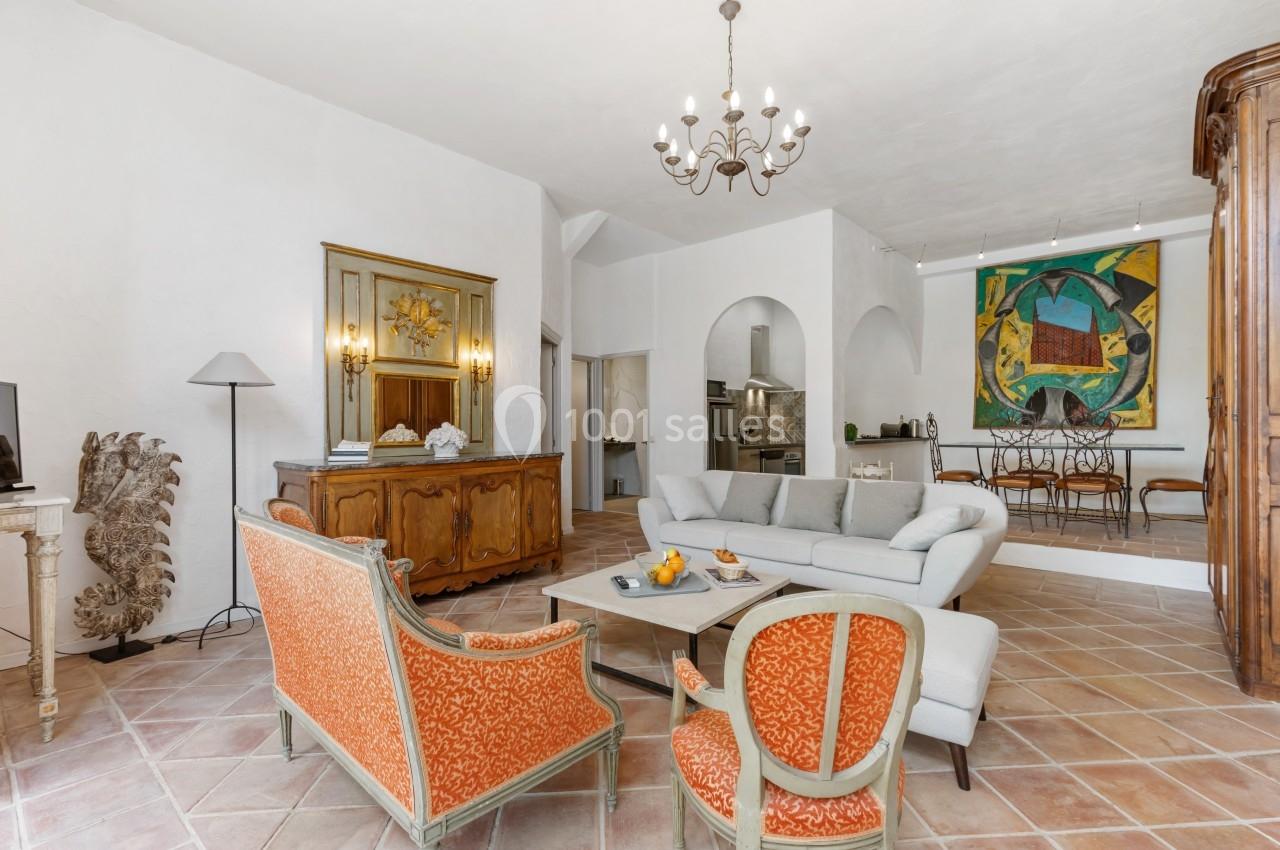 Salon lumineux avec canapé gris, fauteuils orange, table basse, lustre, tableau coloré et mobilier ancien.