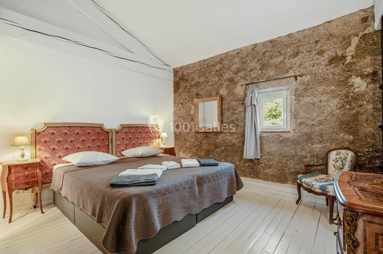 Chambre avec lit double, murs en pierre et bois, sol clair, fenêtre avec rideau gris et mobilier ancien.