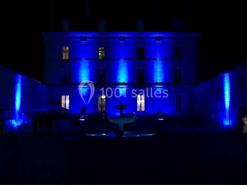 Façade d'un bâtiment éclairé en bleu la nuit, avec une fontaine au premier plan.