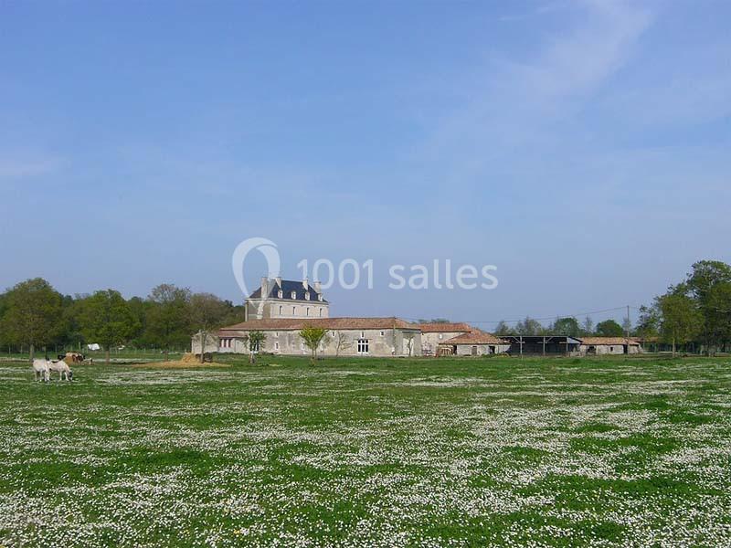 Bâtisse en pierre entourée de prairies fleuries sous un ciel bleu, avec des arbres en arrière-plan.