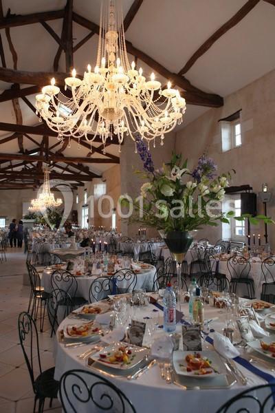 Salle de réception élégante avec tables rondes dressées, chandeliers en cristal et poutres apparentes au plafond.