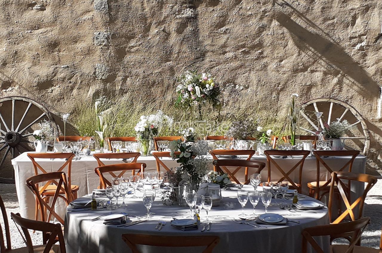 Tables dressées pour un repas en extérieur, avec nappes blanches, chaises en bois et décor floral devant un mur en pierre.