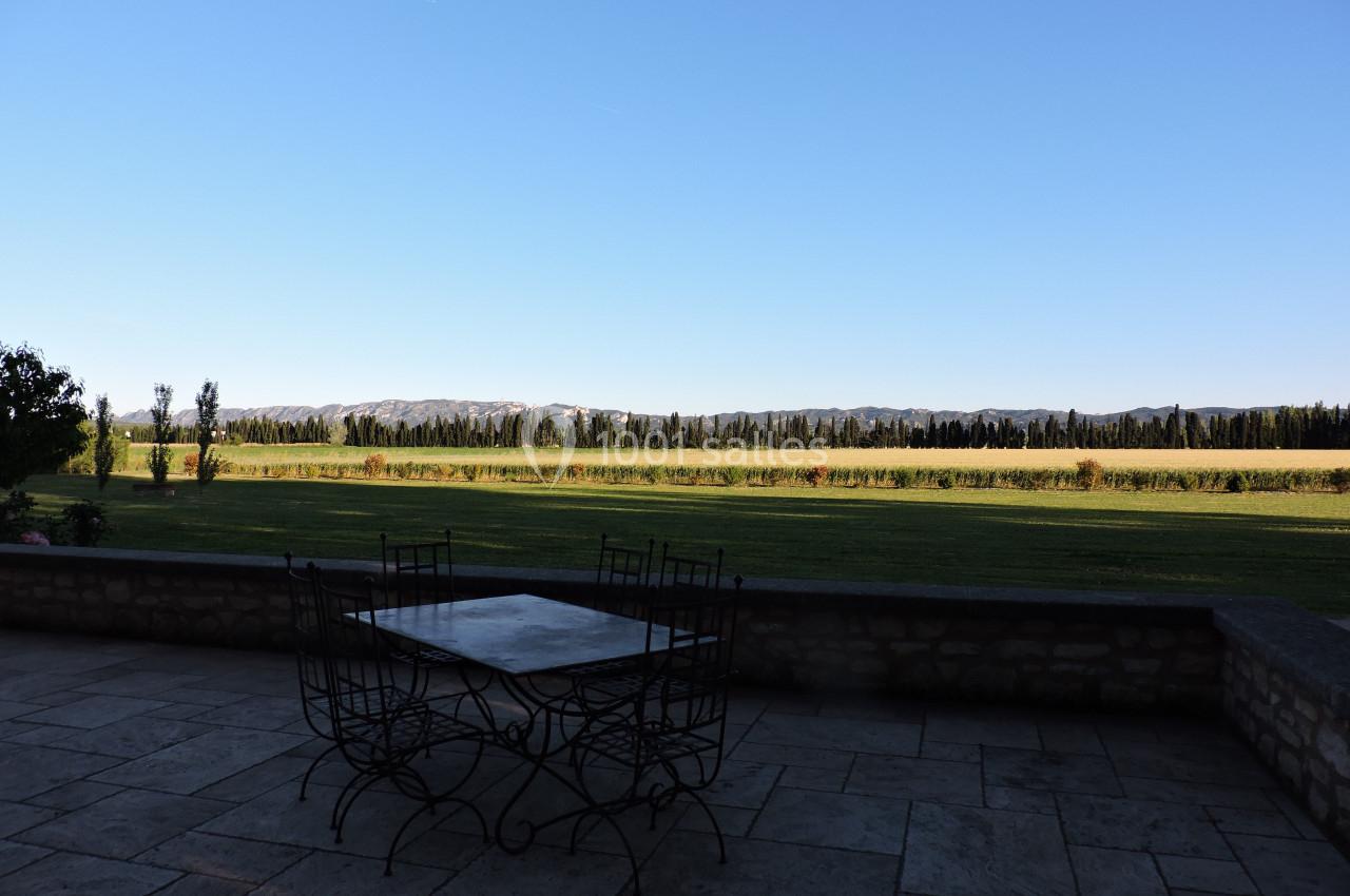 Vue d'une terrasse en pierre avec une table et des chaises en fer forgé, donnant sur un champ et des collines à l'horizon.
