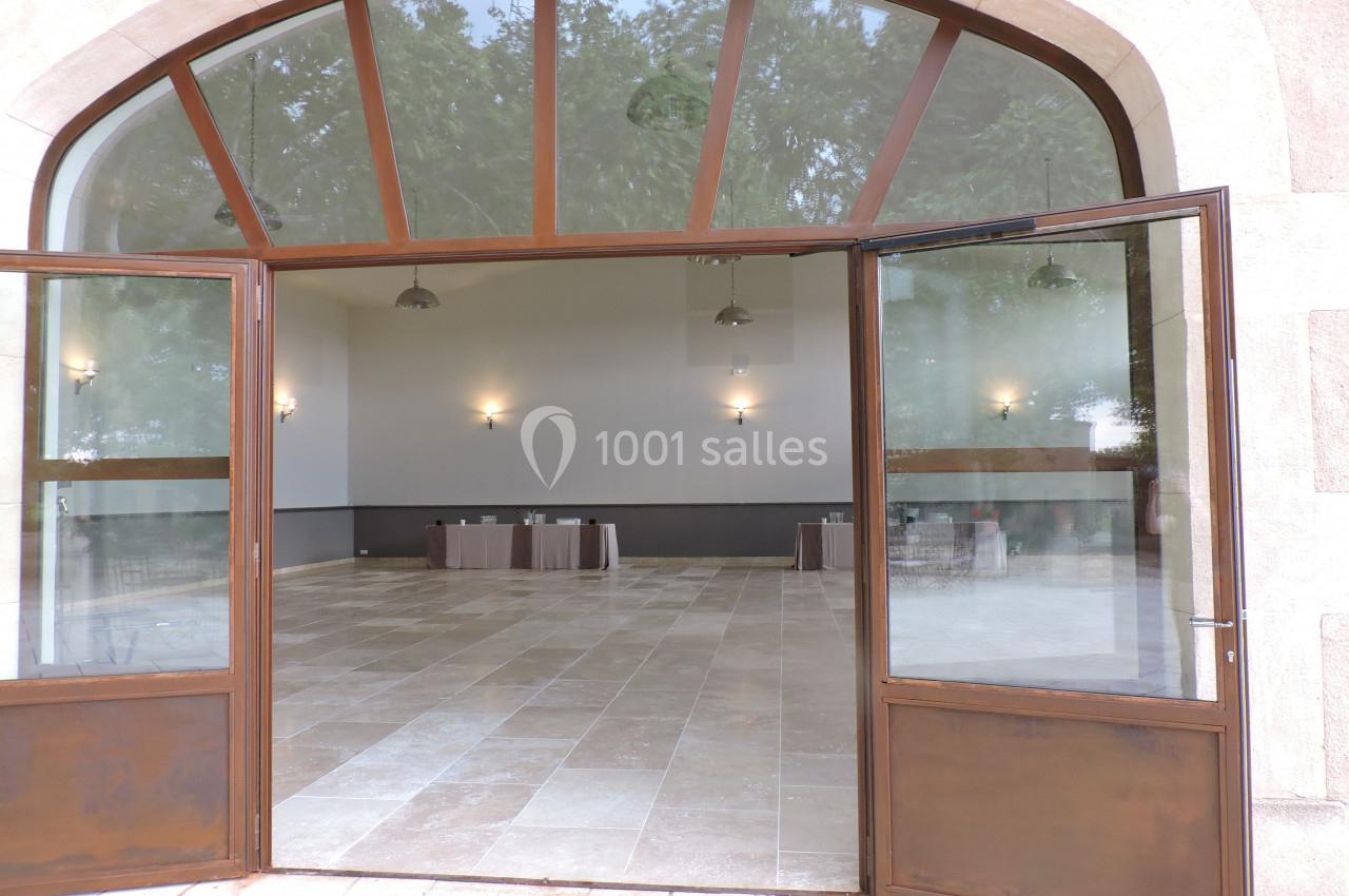 Salle vide avec sol en carrelage beige, murs blancs, grandes baies vitrées et tables couvertes de nappes brunes au fond.