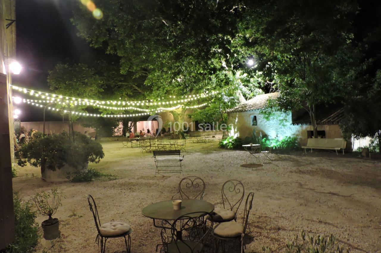 Cour extérieure éclairée par des guirlandes lumineuses, avec des tables et des chaises en métal sous des arbres la nuit.
