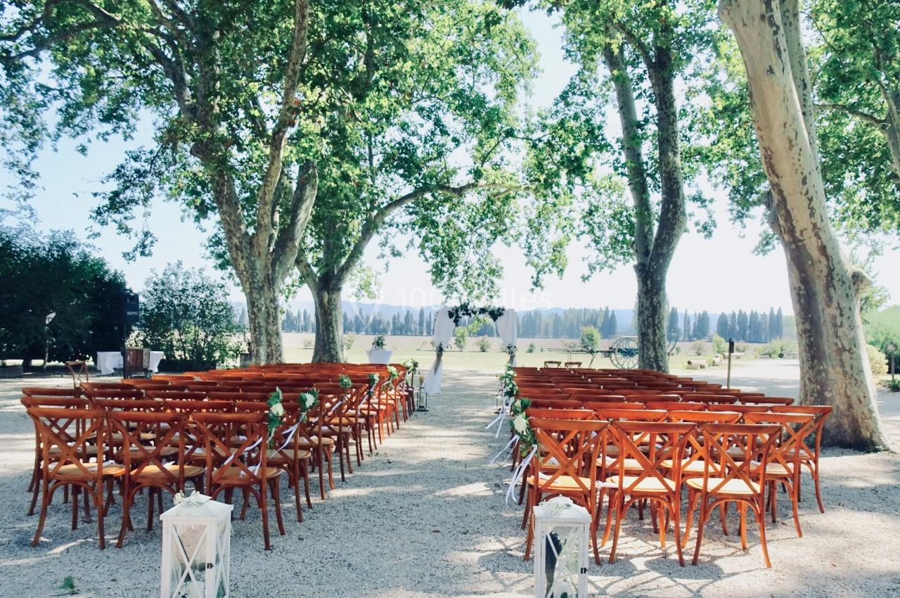 Chaises en bois disposées en rangées sous des arbres, formant une allée menant à une arche décorée pour une cérémonie en…