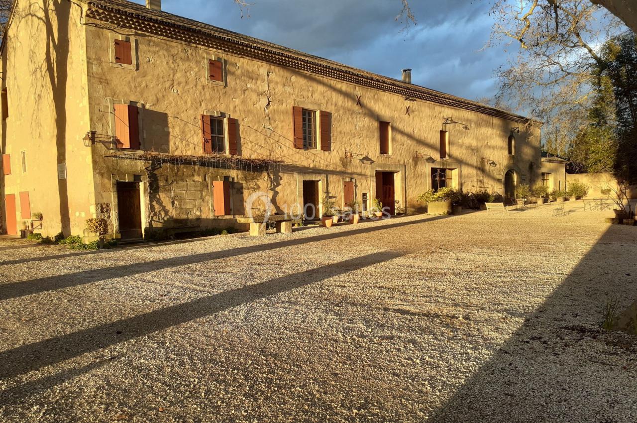 Bâtisse en pierre avec volets rouges, éclairée par une lumière dorée, entourée d'une cour gravillonnée et d'arbres.