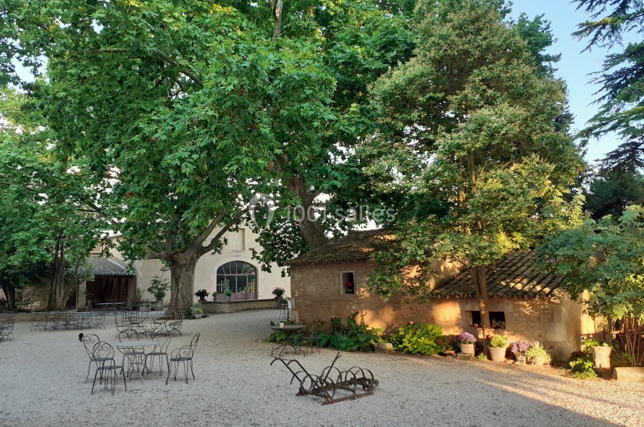 Cour extérieure ombragée avec des arbres, des tables en fer forgé, des bâtiments en pierre et des plantes décoratives.