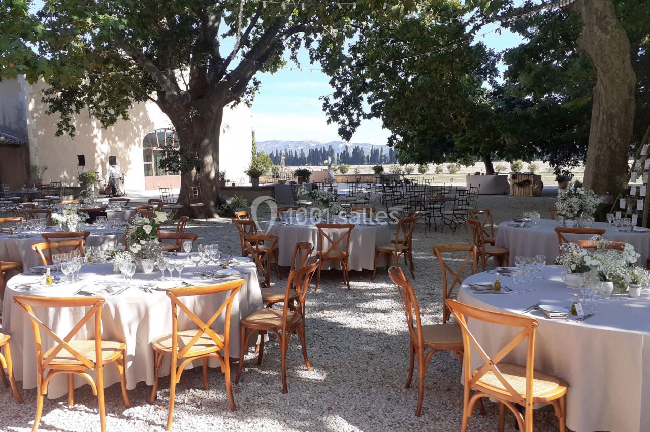 Tables rondes décorées pour un événement en extérieur, disposées sous des arbres dans une cour ensoleillée.