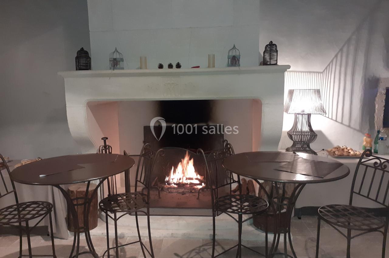 Cheminée allumée dans une pièce avec des tables et chaises en métal, éclairée par une lampe et des bougies.