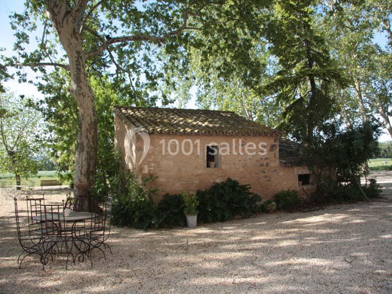 Petite maison en pierre entourée d'arbres, avec une table et des chaises en fer forgé sur un sol gravillonné.