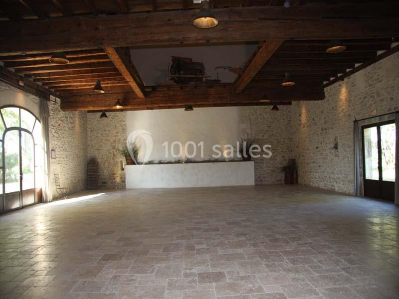 Salle vide avec murs en pierre, sol en carrelage clair, poutres en bois au plafond et grandes ouvertures vitrées.