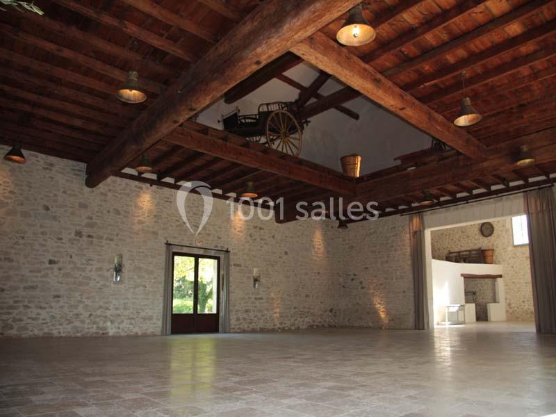 Salle spacieuse avec murs en pierre, poutres en bois apparentes et éclairage suspendu, donnant sur une porte vitrée.