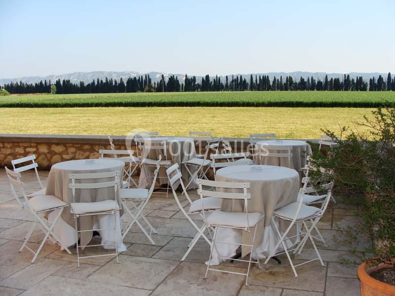 Tables et chaises blanches disposées sur une terrasse en pierre, avec vue sur un champ et des arbres en arrière-plan.