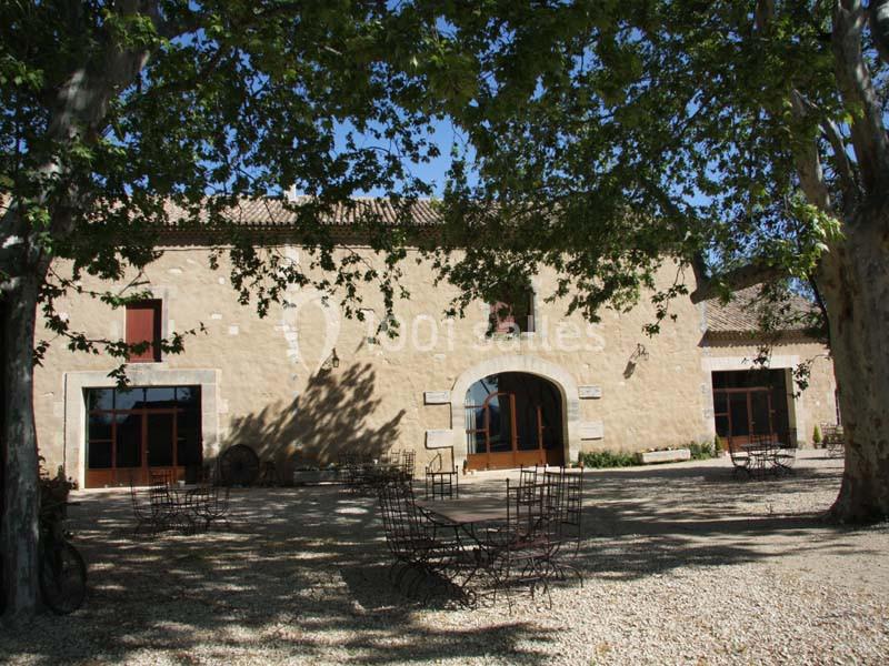 Façade d'un bâtiment en pierre avec des arbres ombrageant une cour équipée de tables et chaises en métal.