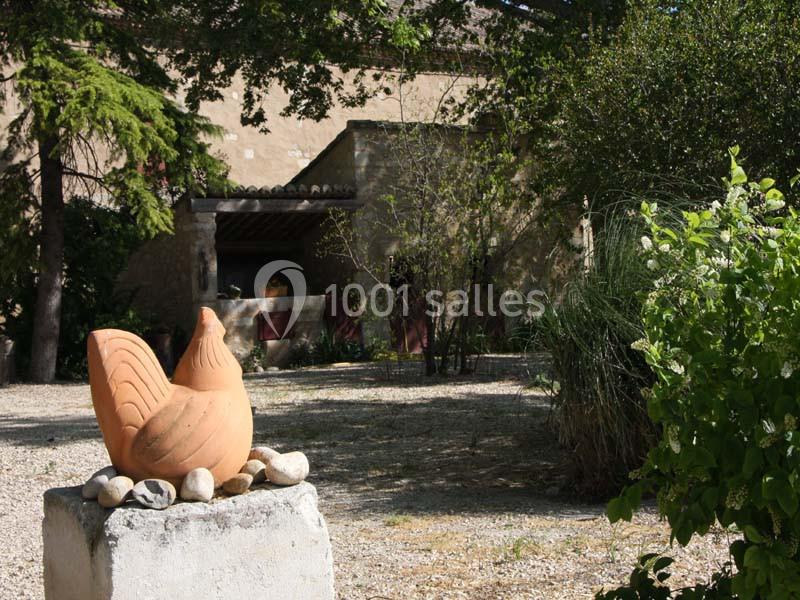 Statue en terre cuite en forme de poule posée sur un socle dans un jardin arboré devant un bâtiment en pierre.