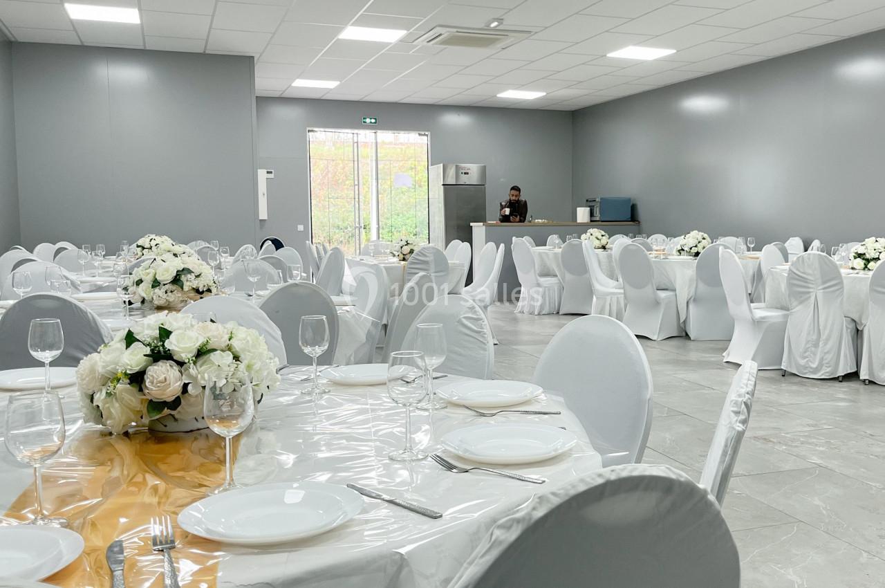 Salle de réception décorée avec des tables rondes, nappes blanches, centres de table fleuris et chaises houssées.