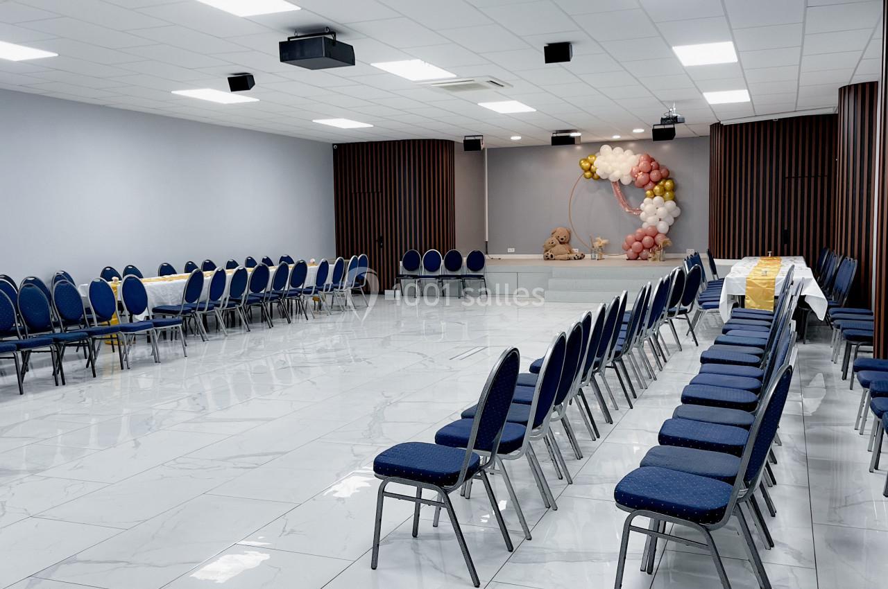 Salle lumineuse avec des chaises bleues disposées en rangées, décorations florales et ballons au fond.