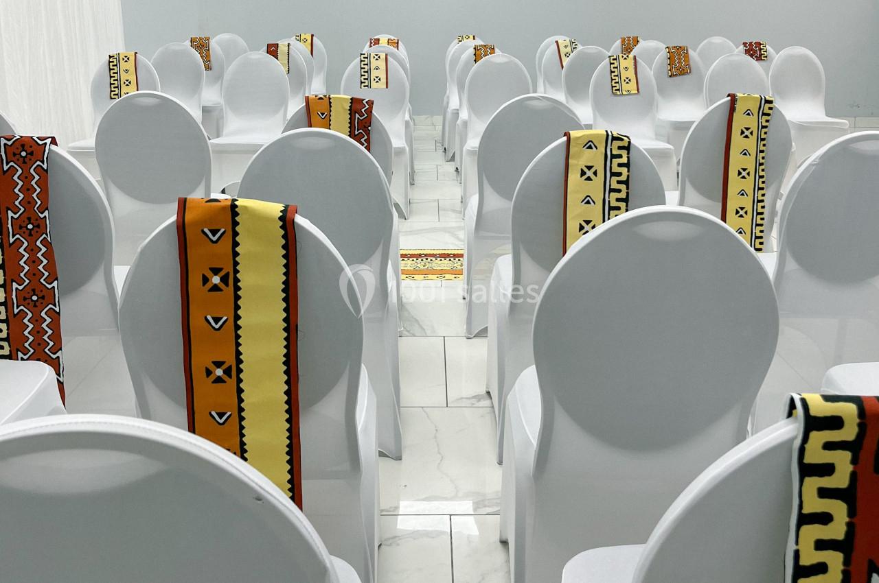 Chaises blanches alignées dans une salle, décorées de tissus colorés à motifs géométriques.