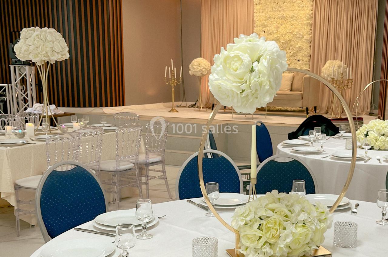 Salle de réception décorée avec des tables dressées, des chaises transparentes et des arrangements floraux blancs.