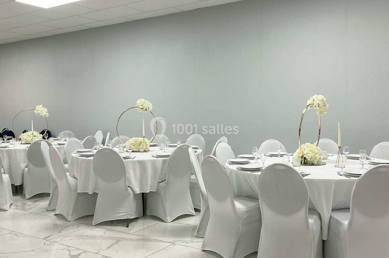 Salle de réception avec tables rondes dressées, nappes blanches, chaises houssées et décorations florales élégantes.