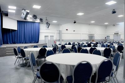 Salle de réception avec tables rondes couvertes de nappes blanches, chaises bleues et une scène avec rideaux bleus.