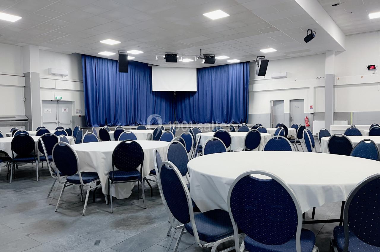 Salle de réception avec des tables rondes couvertes de nappes blanches et des chaises bleues, face à une scène avec rideaux…