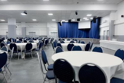 Salle de réception avec tables rondes couvertes de nappes blanches, chaises bleues et une scène avec rideaux bleus.