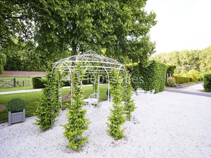 Tonnelle métallique entourée de plantes grimpantes, située sur un sol gravillonné avec des arbres en arrière-plan.
