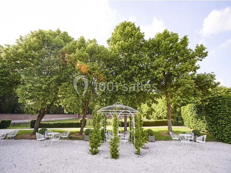 Jardin avec pergola en métal entourée de verdure, tables et chaises blanches sur un sol en gravier.