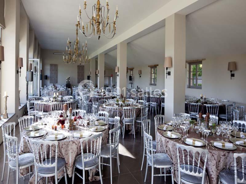 Salle de réception élégante avec tables rondes dressées, chaises blanches et lustres suspendus.