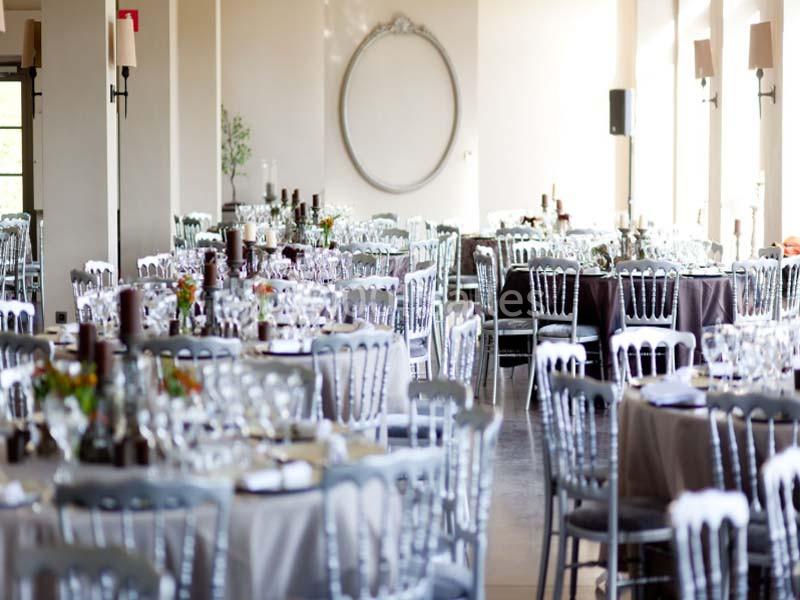 Salle de réception lumineuse avec des tables rondes dressées, entourées de chaises argentées.