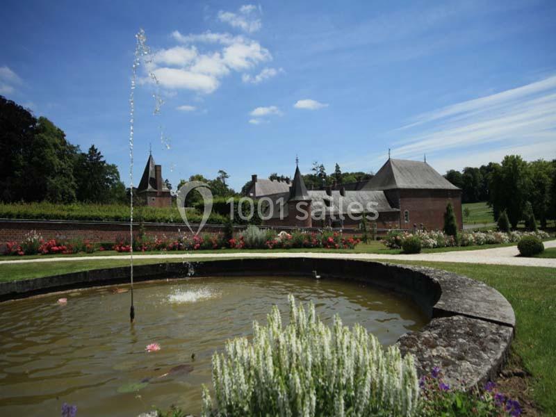 Fontaine entourée de fleurs dans un jardin, avec un bâtiment en briques et des tours en arrière-plan sous un ciel bleu.