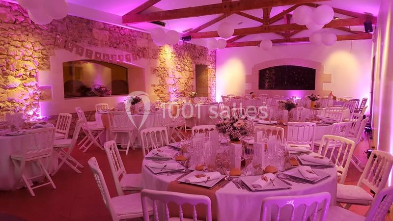 Salle de réception décorée pour un événement, avec tables rondes dressées, chaises blanches et éclairage rose.