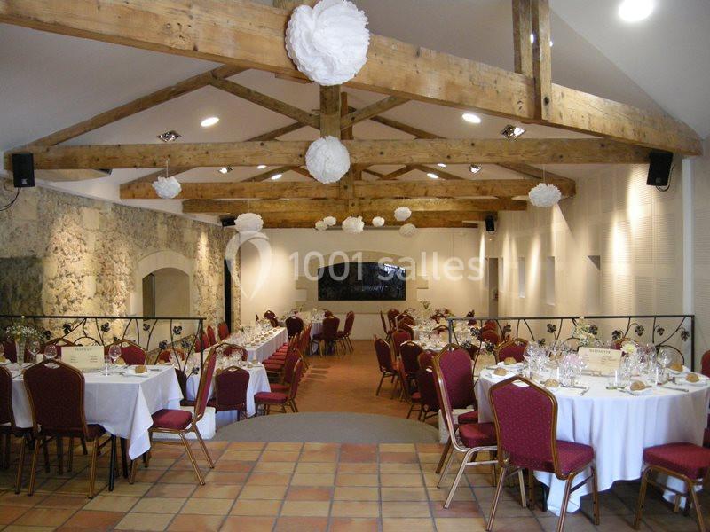Salle de réception avec poutres apparentes, tables rondes dressées et chaises rouges, décorée de suspensions blanches.