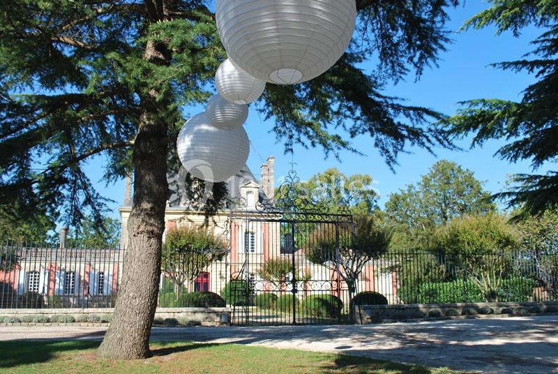 Lanternes blanches suspendues à un arbre devant une grille en fer forgé et un bâtiment ancien entouré de verdure.