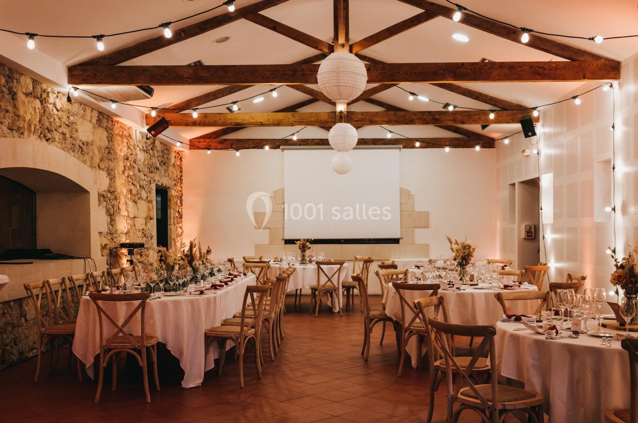 Salle de réception décorée avec des tables rondes dressées, des chaises en bois et des guirlandes lumineuses suspendues.