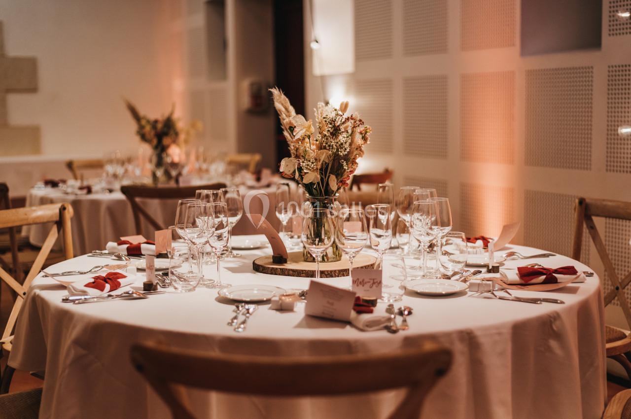 Table ronde élégamment dressée avec vaisselle, verres, serviettes rouges et centre de table floral dans une salle éclairée.