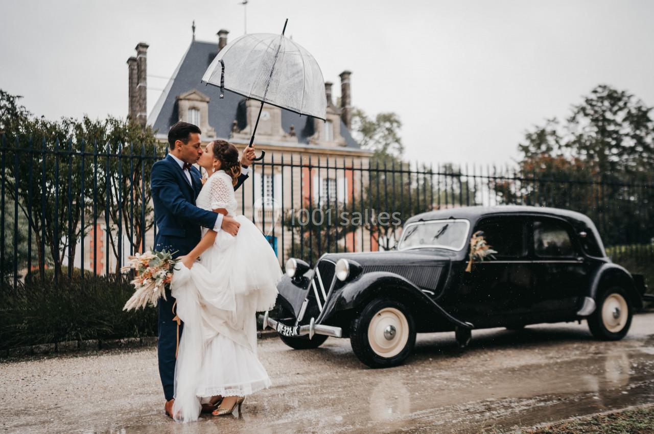 Un couple de mariés sous un parapluie devant une voiture ancienne, avec un manoir en arrière-plan.