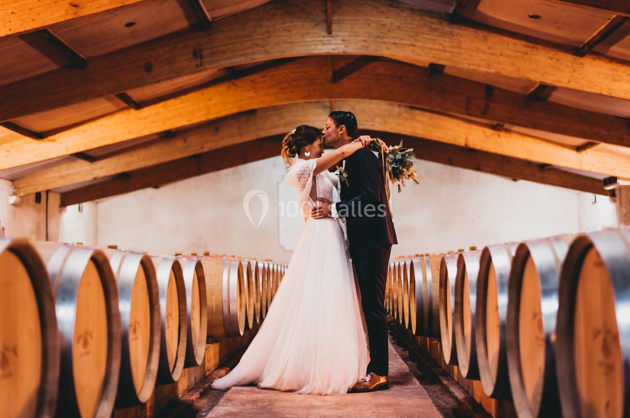 Un couple en tenue de mariage s'embrasse dans une cave à vin entourée de tonneaux en bois.