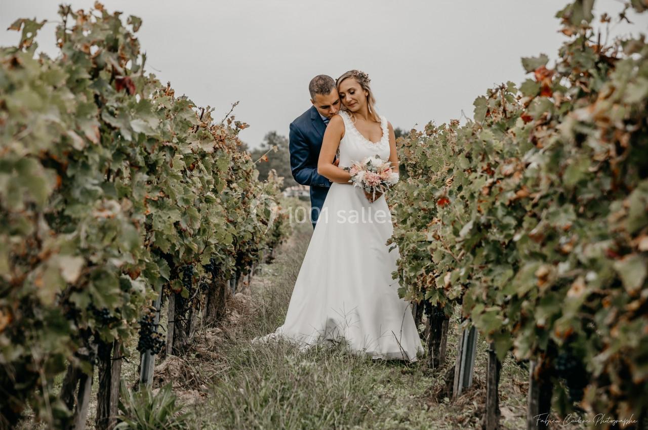 Un couple en tenue de mariage pose dans un vignoble sous un ciel nuageux.