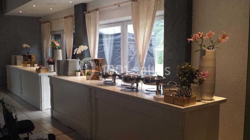 Buffet avec plats chauds sous cloches, vaisselle empilée et décorations florales dans une salle lumineuse.