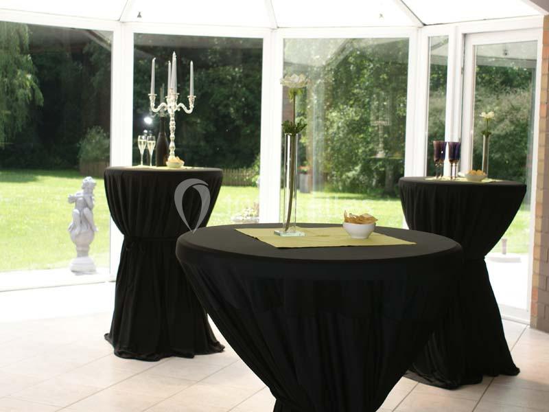 Trois tables hautes noires décorées avec des chandeliers, fleurs et verres, dans une salle lumineuse avec vue sur un jardin.