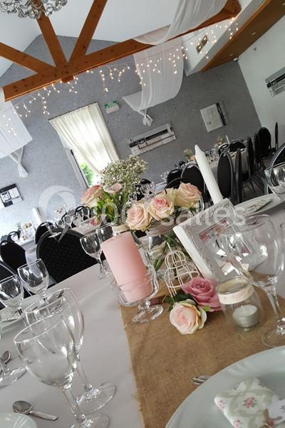 Salle décorée pour un événement avec des tables dressées, des roses, des bougies et des éléments en bois et en toile de jute.