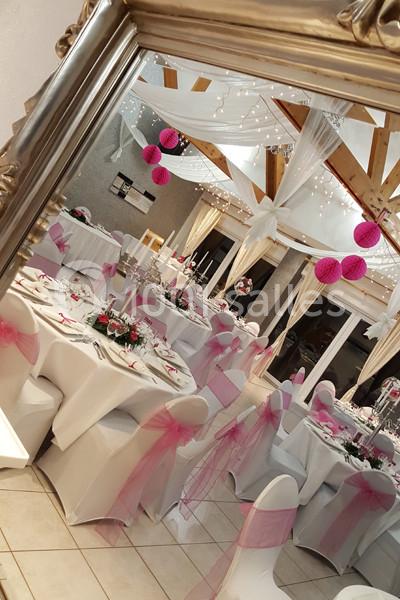 Salle de réception décorée avec des tables rondes, nappes blanches, chaises ornées de rubans roses et guirlandes lumineuses.